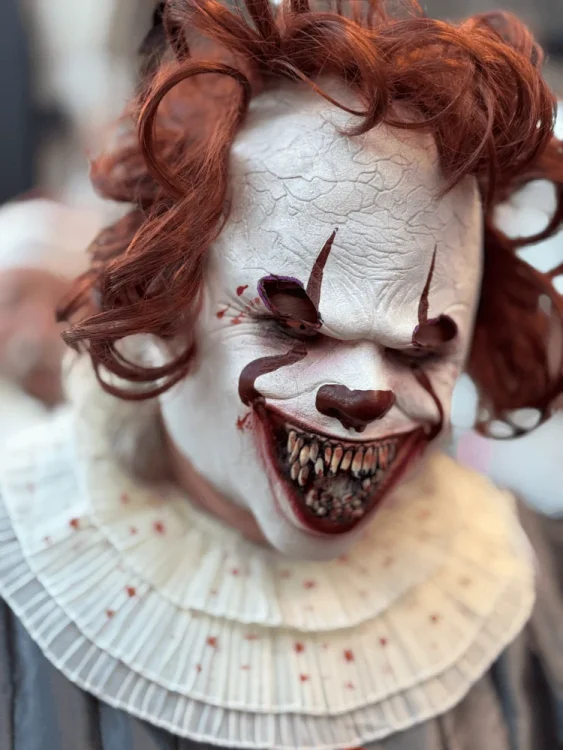 Vosek maske strašljivega klovna z rdečimi lasmi in belim obrazom, na katerem so vidne podrobnosti, kot so razpokanost kože in zobe, ki presegajo človeške značilnosti.