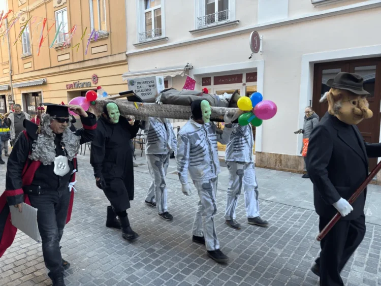 Skupina maskiranih ljudi nosi leseno krsto s pisanimi baloni na pokopu pusta v ulici.
